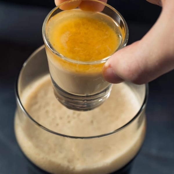 A hand holding a shot glass with a yellow and white layered drink over a larger glass of beer.