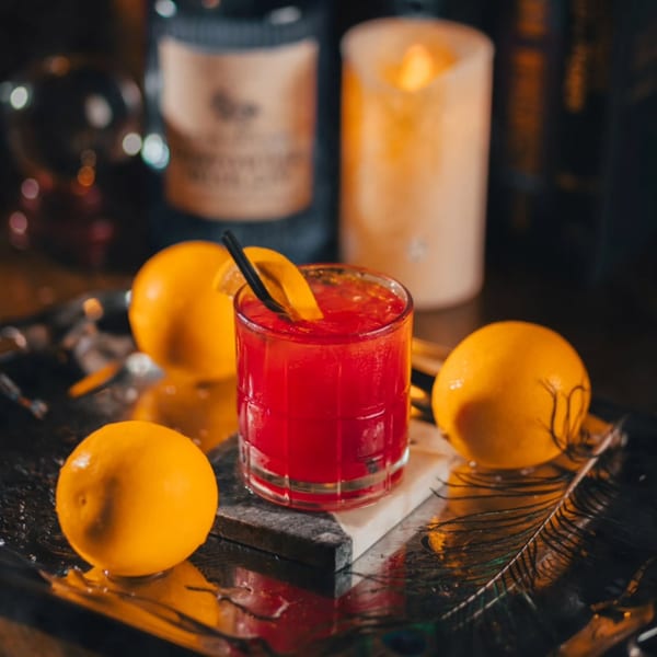 A vibrant red cocktail with a lemon slice and straw, surrounded by whole lemons on a reflective tray.