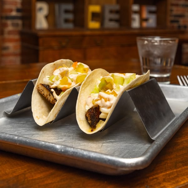 Two Southern-style tacos with chicken, white sauce, and colorful relish on a metal tray.