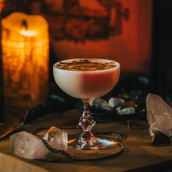 A creamy cocktail with cinnamon dusting sits on a wooden table next to a glowing candle and crystals.