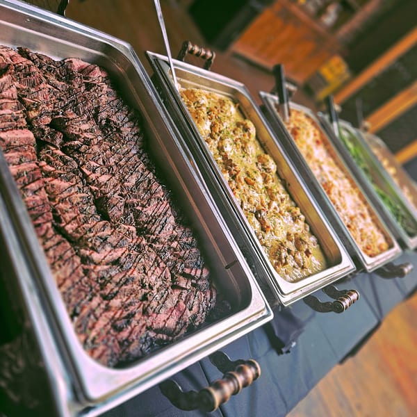 Buffet line with sliced grilled steak, creamy chicken, and other southern dishes in chafing dishes.