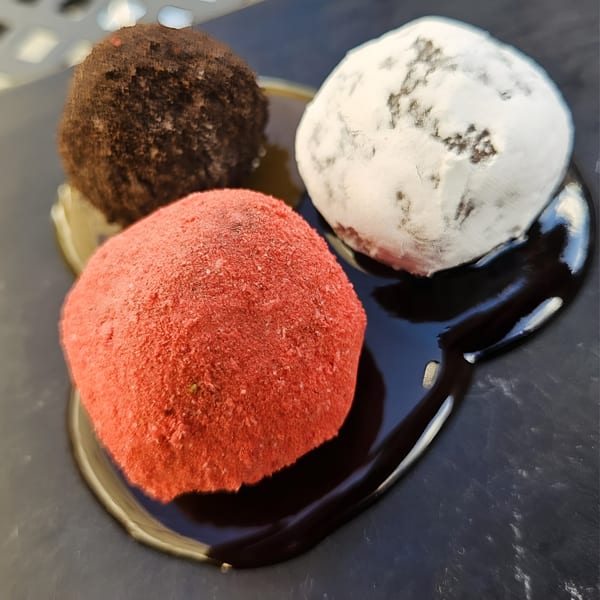 Three colorful dessert balls, red, brown, and white, on a dark plate with chocolate sauce.