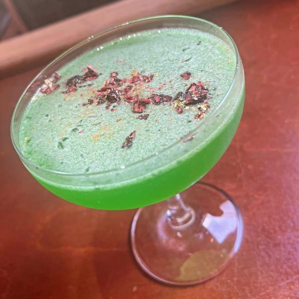 Vibrant green cocktail with a frothy top, garnished with red and gold flakes, on a wooden table.