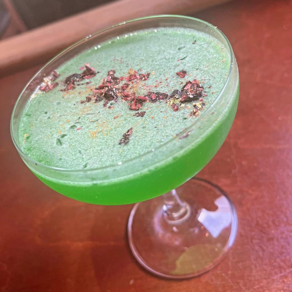Vibrant green cocktail with a frothy top, garnished with red and gold flakes, on a wooden table.