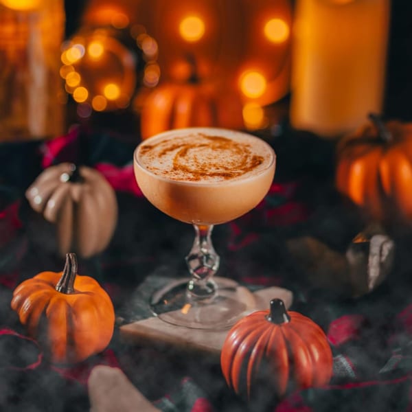 A pumpkin spice cocktail with cinnamon swirl, surrounded by decorative pumpkins and warm lights.