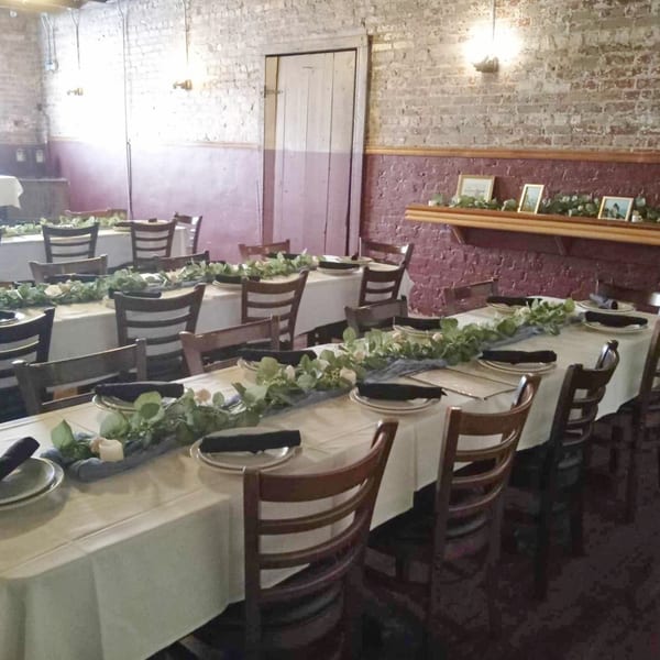 Long dining tables with white tablecloths, place settings, and green garland in a rustic room.